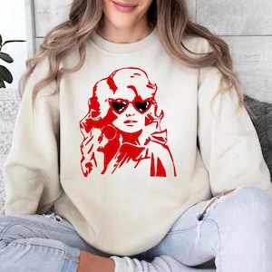 May include: A cream-colored sweatshirt with a red graphic of a woman with blonde hair wearing heart-shaped sunglasses. The woman is looking to the right.