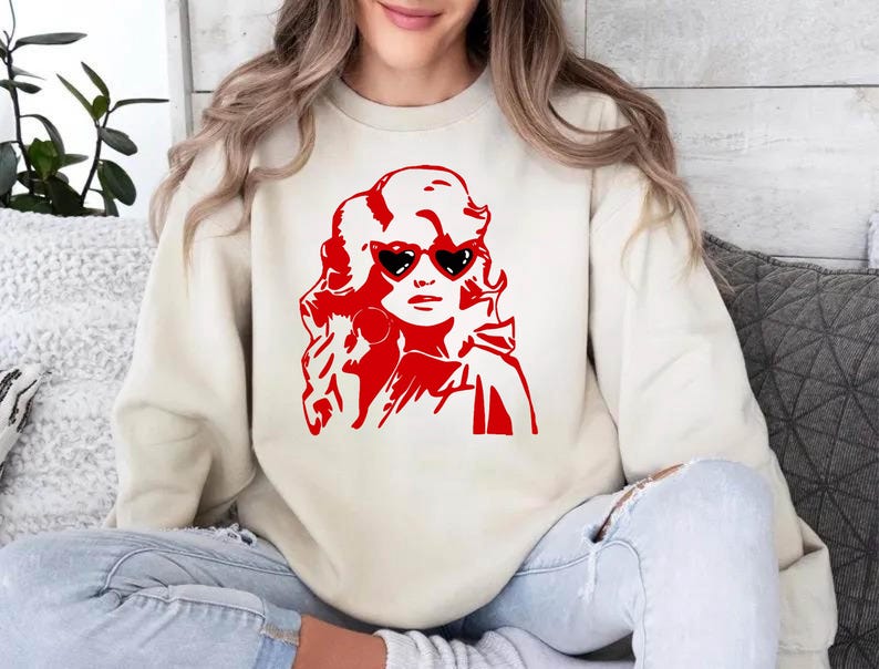 May include: A cream-colored sweatshirt with a red graphic of a woman with blonde hair wearing heart-shaped sunglasses. The woman is looking to the right.