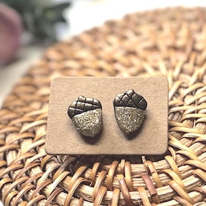 May include: A pair of acorn-shaped stud earrings featuring dark brown textured caps and shimmering gold glitter bases. The earrings are attached to a rectangular brown card, displaying a detailed design that mimics the appearance of natural acorns.