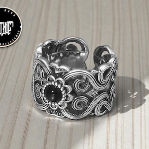 May include: A silver-toned adjustable ring with an ornate, gothic design. The ring features a central black stone surrounded by a floral pattern, with swirling details and the word "GOTHIC" in a circular logo.