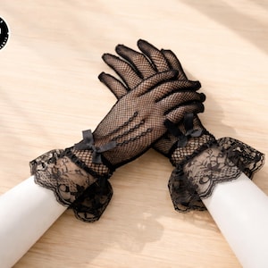 Puede incluir: Guantes negros de red con puños de encaje y pequeños lazos. Los guantes se exhiben sobre formas blancas sobre un fondo de madera clara. Los guantes tienen una estética gótica.