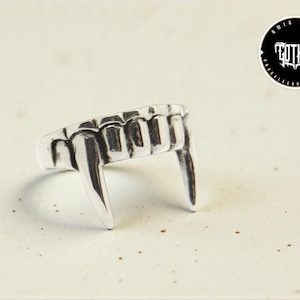 May include: A silver-colored ring shaped like vampire fangs. The ring features two prominent fangs and several smaller teeth. The ring is on a speckled, off-white surface. A black and white circular logo with the word "Gothic" is in the background.