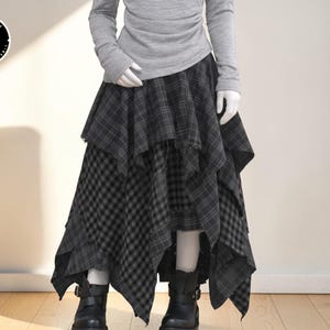 Puede incluir: Una falda asimétrica de varias capas en tonos grises y negros con estampados de cuadros y cuadros. La falda se combina con una camiseta gris de manga larga y botas negras. La imagen presenta una estética de moda gótica.