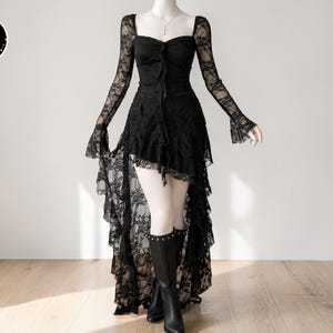 May include: Black lace dress with long sleeves and a high-low hemline. The dress has a square neckline and a fitted bodice. The sleeves are fitted with flared cuffs. The dress is styled with black boots. A logo is in the upper left corner.
