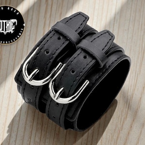 May include: Black leather wrist cuff with two straps and silver buckles. The cuff features a stacked design and is fastened with adjustable buckles. The image also includes a circular logo with the word "GOTHIC".