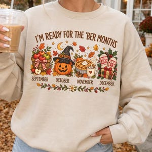 May include: A beige crewneck sweatshirt featuring the text I'M READY FOR THE 'BER MONTHS. The graphic includes a basket of apples, a jack-o'-lantern wearing a witch hat, a pumpkin pie, and a gift box with mittens, labeled September to December.