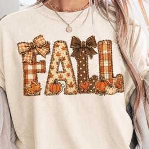 Puede incluir: Una sudadera de cuello redondo color crema con la palabra FALL en letras grandes y decorativas. Cada letra está rellena con patrones otoñales que incluyen cuadros naranjas, hojas de arce y lunares, decorada con lazos marrones y calabazas.