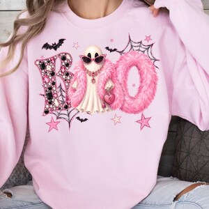 Puede incluir: Una sudadera de cuello redondo rosa claro con un estampado gráfico de la palabra BOO. La letra B está decorada con gemas negras y rosas, mientras que la letra O está cubierta de piel sintética rosa. Un fantasma lleva gafas de sol rosas.