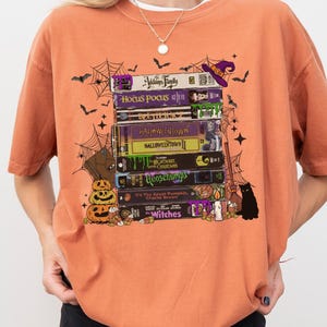 Puede incluir: Camiseta naranja con un diseño de cintas VHS apiladas con temática de Halloween, incluyendo "Hocus Pocus" y "Witches". El diseño también presenta murciélagos, telarañas, un sombrero de bruja, un gato negro y calabazas. La camiseta tiene un ajuste holgado y extragrande.