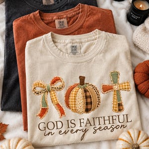 May include: Three crewneck t-shirts in black, burnt orange, and cream. The cream shirt features a graphic of a bow, a pumpkin, and a cross in plaid and leopard patterns, with the text "GOD IS FAITHFUL in every season" printed below.