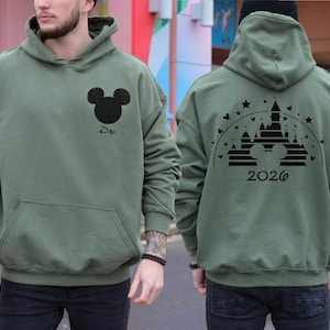 Puede incluir: Sudadera con capucha verde salvia con un gr&aacute;fico de la cabeza de Mickey Mouse en negro y la palabra "Dad" en la parte delantera. La parte trasera presenta un dise&ntilde;o de castillo con el a&ntilde;o "2026". La sudadera tiene un bolsillo delantero y una capucha.