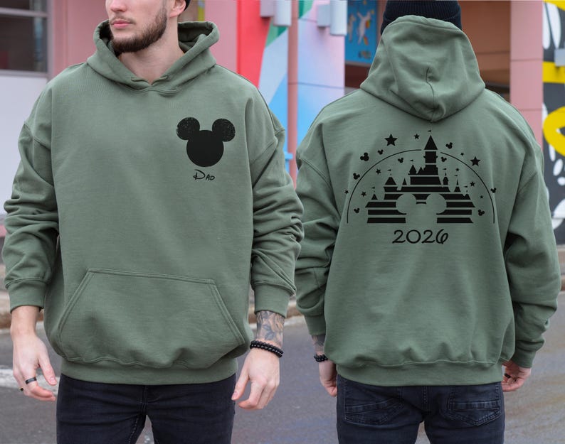 Puede incluir: Sudadera con capucha verde salvia con un gr&aacute;fico de la cabeza de Mickey Mouse en negro y la palabra "Dad" en la parte delantera. La parte trasera presenta un dise&ntilde;o de castillo con el a&ntilde;o "2026". La sudadera tiene un bolsillo delantero y una capucha.