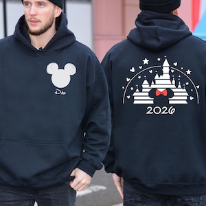 Puede incluir: Sudadera con capucha azul marino con un dise&ntilde;o de cabeza de Mickey Mouse blanca y la palabra "Dad" debajo. La parte trasera muestra un dise&ntilde;o de castillo blanco con una pajarita roja y el a&ntilde;o "2026".
