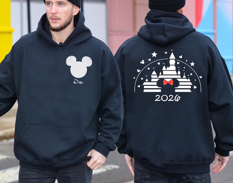 Puede incluir: Sudadera con capucha azul marino con un dise&ntilde;o de cabeza de Mickey Mouse blanca y la palabra "Dad" debajo. La parte trasera muestra un dise&ntilde;o de castillo blanco con una pajarita roja y el a&ntilde;o "2026".