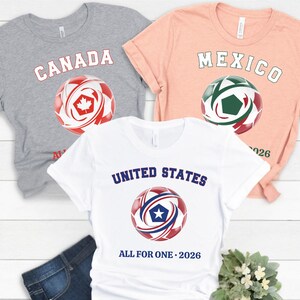 May include: Three short-sleeved t-shirts with soccer ball graphics. The shirts are gray, peach, and white. The gray shirt says "CANADA" with a red and white soccer ball. The peach shirt says "MEXICO" with a green and red soccer ball. The white shirt says "UNITED STATES" with a red and blue soccer ball.
