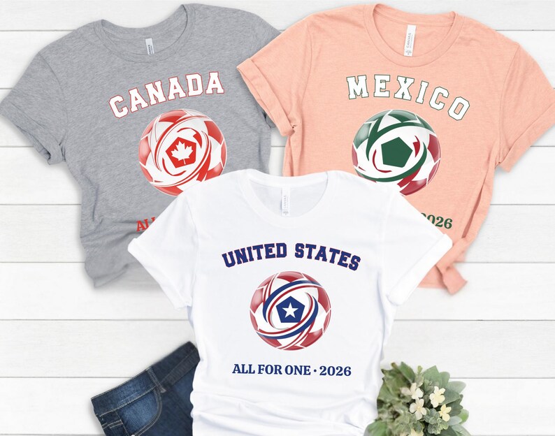 May include: Three short-sleeved t-shirts with soccer ball graphics. The shirts are gray, peach, and white. The gray shirt says "CANADA" with a red and white soccer ball. The peach shirt says "MEXICO" with a green and red soccer ball. The white shirt says "UNITED STATES" with a red and blue soccer ball.