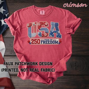 Puede incluir: Camiseta de color carmesí con un diseño de patchwork falso. La palabra "USA" se muestra en un diseño patriótico, con el texto "250 YEAR OF FREEDOM" debajo. La camiseta está hecha de un material suave.