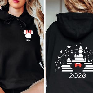 Puede incluir: Sudadera con capucha negra con dise&ntilde;o de Minnie Mouse. La parte delantera muestra una cabeza de Minnie Mouse con un lazo rojo y la palabra "Mom". La parte trasera presenta una silueta de castillo, estrellas y el a&ntilde;o "2026".