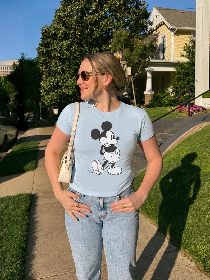 May include: A light blue t-shirt featuring a black and white graphic of Mickey Mouse. The shirt is paired with light wash blue jeans. A white purse is worn over the shoulder. The person is wearing sunglasses and rings.