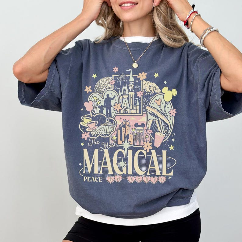 May include: A slate blue short-sleeve t-shirt featuring a graphic print of a castle, park icons, and flowers in pastel colors. The text reads The Most MAGICAL PLACE ON EARTH, with small heart shapes surrounding the words at the bottom of the design.