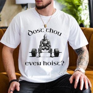 May include: White t-shirt with a black and white illustration of a knight lifting a barbell. The text "Dost thou even hoist?" is arched above and below the knight image. The shirt is made of a soft material.