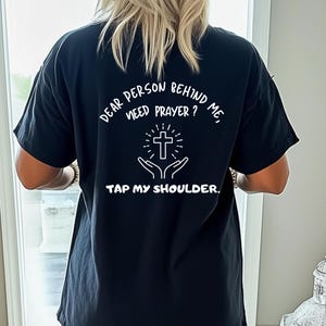 May include: Black t-shirt with white text that reads "Dear person behind me, need prayer? Tap my shoulder."  The text is surrounded by a white cross and hands holding the cross.