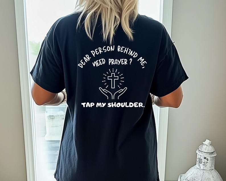 May include: Black t-shirt with white text that reads "Dear person behind me, need prayer? Tap my shoulder."  The text is surrounded by a white cross and hands holding the cross.