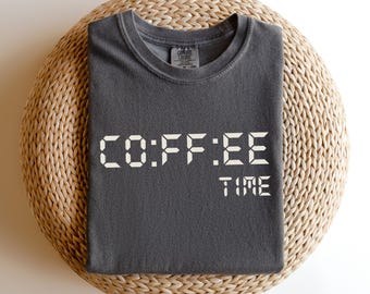May include: Dark grey t-shirt with the words "COFFEE TIME" in a digital font. The shirt is folded on a round, woven, light brown surface. The t-shirt has a crew neck and short sleeves.