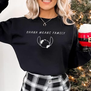 May include: Black sweatshirt with the text "OHANA MEANS FAMILY" and a white outline of a character. A person is holding a red and white Santa-themed mug. The person is wearing a silver necklace and a black and white plaid skirt.