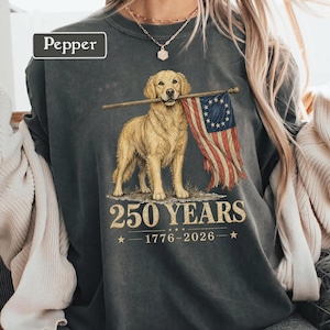May include: A charcoal gray t-shirt featuring a golden retriever holding a wooden pole with a historical American flag. Below the dog, the text reads 250 YEARS 1776-2026. The word Pepper is printed in a small box in the upper left corner of the shirt.
