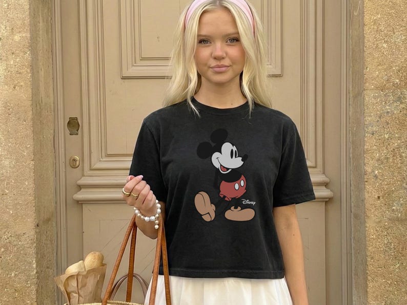以下が含まれることがあります： クラシックなミッキーマウスのグラフィックが特徴の黒いTシャツ。Tシャツはクルーネックと半袖です。ピンクのヘアバンドを着用し、茶色のレザートートバッグを持っています。