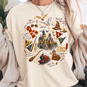 May include: A cream-colored crewneck t-shirt featuring a detailed graphic print of a castle, a steam train, a broomstick, a scarf, a sorting hat, and various magical icons. The text includes phrases like After all this time, Always, and Mischief Managed.