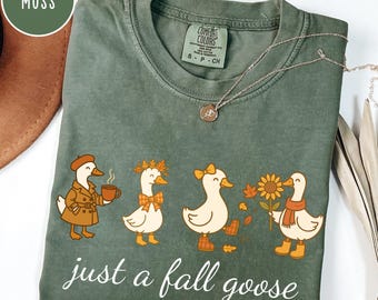 Puede incluir: Camiseta Comfort Colors verde musgo con una ilustración caprichosa de cuatro gansos vestidos con atuendos otoñales. Un ganso sostiene una taza de café, otro lleva una corona de hojas. El texto "just a fall goose" está impreso debajo.