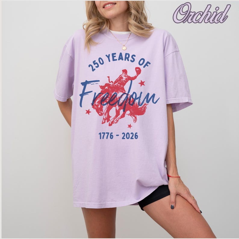 May include: A light purple t-shirt with the text "250 Years of Freedom" and the years 1776-2026. The graphic features a red silhouette of a person riding a horse. The shirt is a relaxed fit.