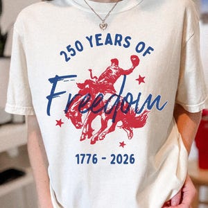 May include: Cream-colored t-shirt with a red and blue graphic celebrating 250 years of freedom. The design features a cowboy riding a bucking bronco, the text "250 Years of Freedom" and the years "1776-2026".