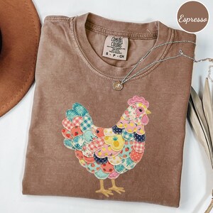 Op de afbeelding: Espresso-kleurig T-shirt met een kleurrijk patchwork kippenontwerp. De kip heeft verschillende patronen en kleuren, waaronder blauw, rood, geel en groen. Een zilveren ketting met een kleine hanger is over het shirt gedrapeerd. Het shirt heeft een Comfort Colors-label.