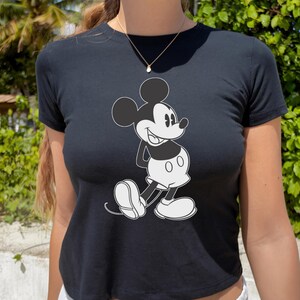 May include: Black crop top featuring a classic Mickey Mouse graphic in black and white. The cartoon character is depicted in a side pose with a cheerful expression. The top has short sleeves and a round neckline, perfect for casual wear.