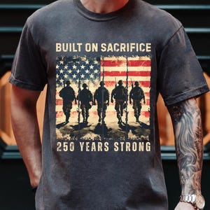 May include: Dark gray t-shirt featuring an American flag design with soldiers and the text "BUILT ON SACRIFICE" and "250 YEARS STRONG". The graphic is distressed, giving a vintage look.