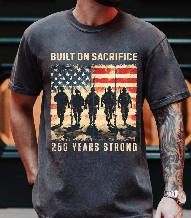 May include: Dark gray t-shirt featuring an American flag design with soldiers and the text "BUILT ON SACRIFICE" and "250 YEARS STRONG". The graphic is distressed, giving a vintage look.