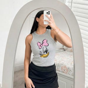 Op de afbeelding: Een grijze tanktop met een cartoonafbeelding van een roze strik, wimpers en een gele snavel. De tanktop wordt gecombineerd met een zwarte rok. De foto is genomen voor een spiegel.