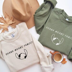 May include: Two sweatshirts, one cream and one olive green, with the text "OHANA MEANS FAMILY" and a cartoon character outline. A brown hat and a fall leaf are also in the image.
