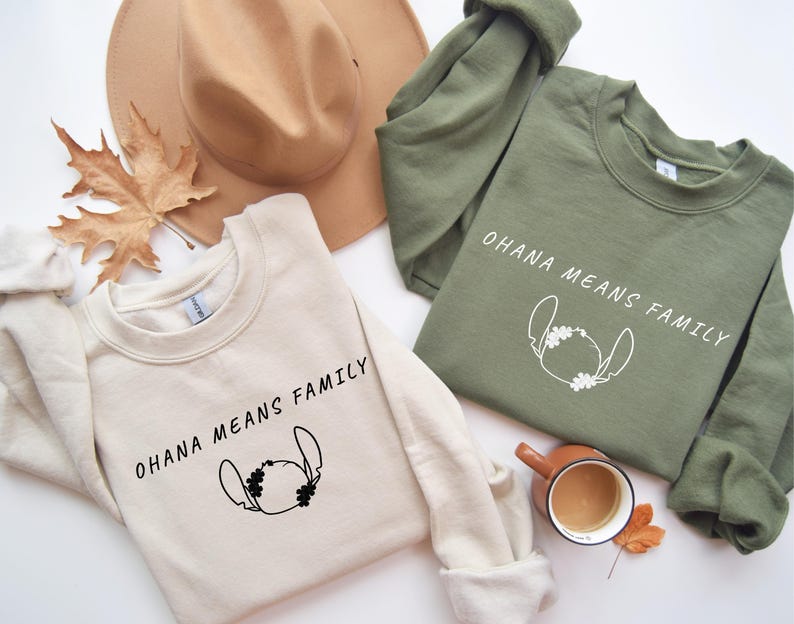 May include: Two sweatshirts, one cream and one olive green, with the text "OHANA MEANS FAMILY" and a cartoon character outline. A brown hat and a fall leaf are also in the image.
