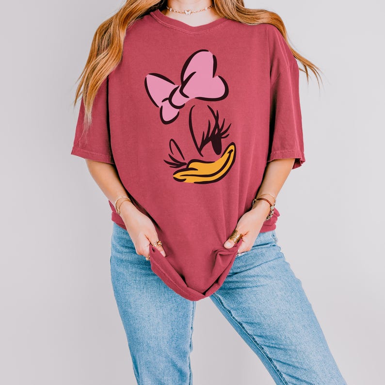 May include: A maroon t-shirt featuring a cartoon graphic of a duck's face with a pink bow, eyelashes, and a yellow beak. The shirt has short sleeves and a relaxed fit. The model is wearing blue jeans.