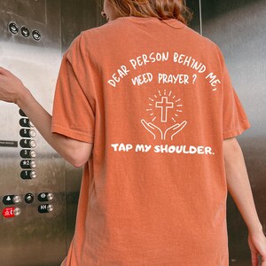 May include: A rust-colored t-shirt with the text "Dear person behind me, need prayer? Tap my shoulder." and a white outline of a cross and hands in a praying position.