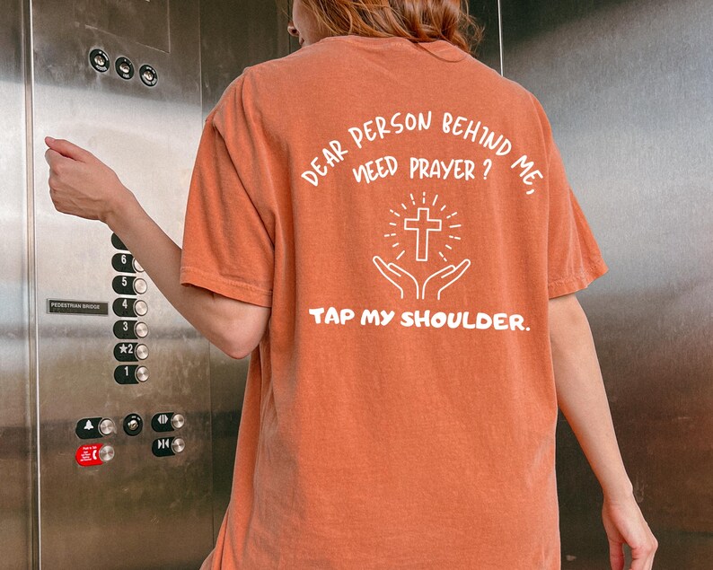 May include: A rust-colored t-shirt with the text "Dear person behind me, need prayer? Tap my shoulder." and a white outline of a cross and hands in a praying position.