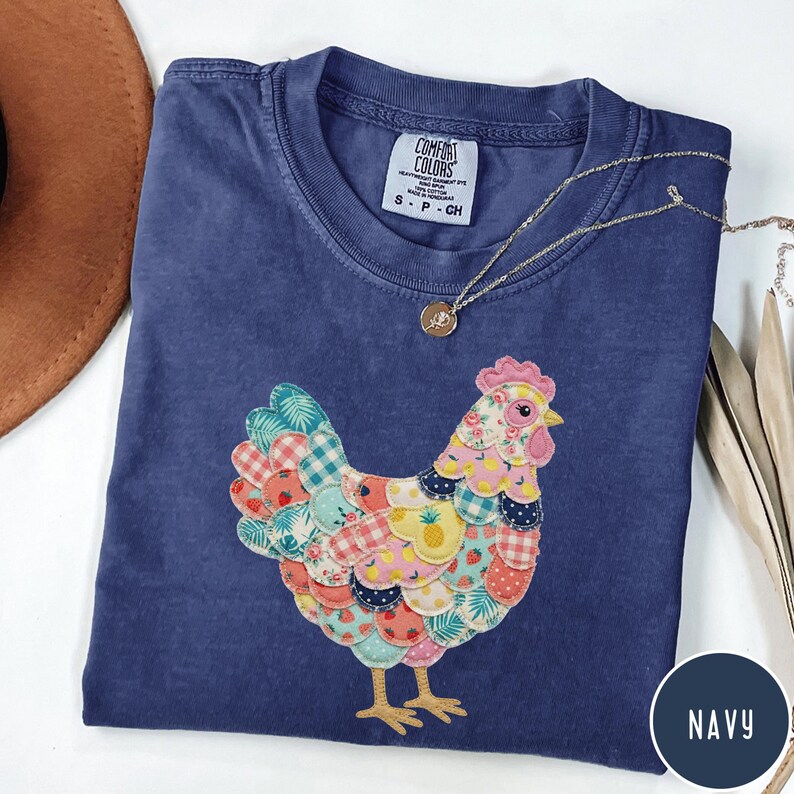 Op de afbeelding: Marineblauw T-shirt met een kleurrijk patchwork kippenontwerp. De kip heeft verschillende patronen en kleuren, waaronder roze, geel en blauw. Een gouden ketting met een hanger is over het shirt gedrapeerd. Het shirt heeft het Comfort Colors label.