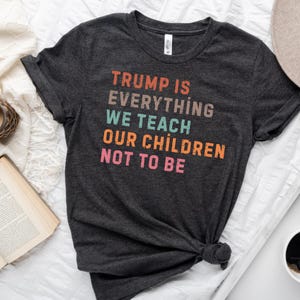 Puede incluir: Camiseta gris oscuro con el texto "TRUMP IS EVERYTHING WE TEACH OUR CHILDREN NOT TO BE" en letras de colores. La camiseta está anudada en la parte inferior.