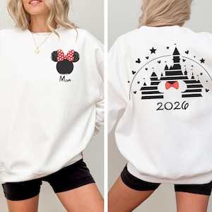 Puede incluir: Sudadera blanca con un dise&ntilde;o de Minnie Mouse, un lazo rojo de lunares y la palabra "Mom". La parte trasera muestra un gr&aacute;fico de castillo con un lazo y el a&ntilde;o "2026".