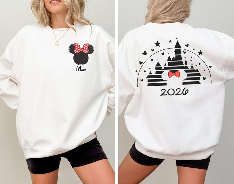 Puede incluir: Sudadera blanca con un dise&ntilde;o de Minnie Mouse, un lazo rojo de lunares y la palabra "Mom". La parte trasera muestra un gr&aacute;fico de castillo con un lazo y el a&ntilde;o "2026".