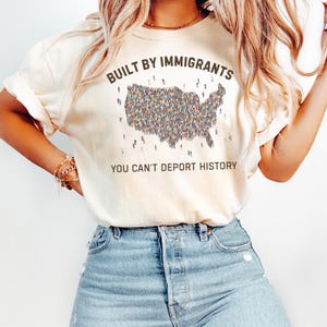 May include: Cream-colored t-shirt with the text "BUILT BY IMMIGRANTS" above a graphic of the United States filled with tiny figures. Below the graphic, the text reads "YOU CAN'T DEPORT HISTORY". The shirt is paired with blue jeans.